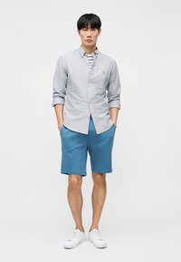 Άνδρας που φοράει ανοιχτό γκρι πουκάμισο με κουμπιά πάνω από ριγέ μπλουζάκι, μπλε σορτς και λευκά αθλητικά παπούτσια, όρθιος με τα χέρια στις τσέπες.