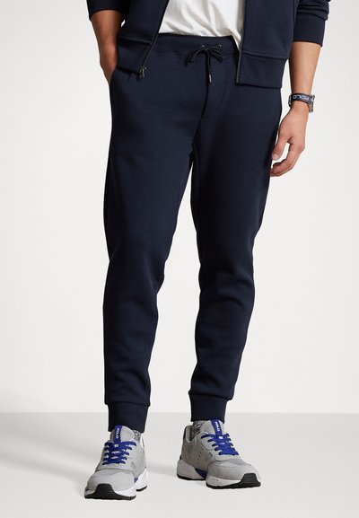 Marineblaue Sweatpants aus weichem Stoff, mit einem elastischen Bund und einer Kordel zum Verstellen, schmal zulaufenden Beinen und Seitentaschen. Kombiniert mit grauen Sneakers.