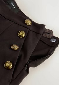 Detailní záběr na tmavohnědý oděv se zdobnými zlatými knoflíky, černým zipem částečně otevřeným a průhledným plastovým drukem na pase.