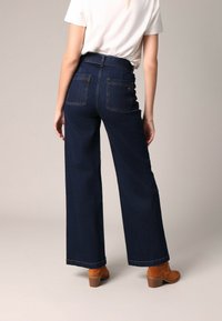 Donkerblauwe wijde jeans met een hoge taille, twee achterzakken en zichtbare stiksels. Gepaard met bruine enkellaarsjes met hak.