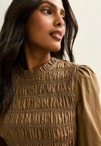 Donna con lunghi capelli scuri che indossa una blusa marrone a collo alto con arricciature, che guarda leggermente in alto e di lato contro uno sfondo chiaro.