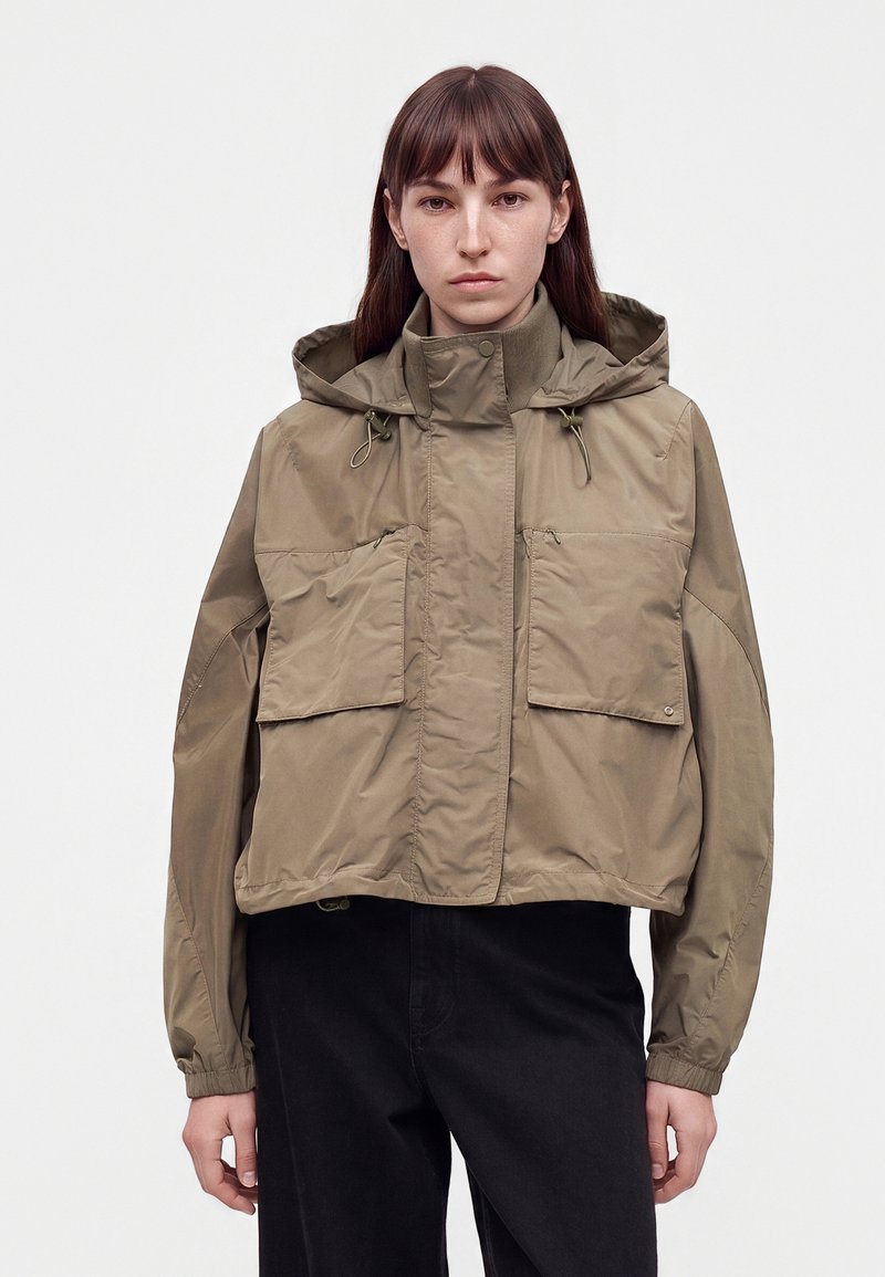 Femme portant une veste à capuche ample vert olive avec de grandes poches avant et un pantalon noir, debout devant un fond blanc uni.