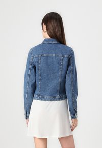 Jeansjacka i ljusblått, med klassisk krage, två bakfickor och knappade ärmar, visad på en modell som bär en vit kjol.