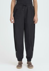 Osoba u crnim jogger hlačama s elastičnim pojasom i bočnim patentnim zatvaračima te smeškim Adidas tenisicama na jednostavnoj svijetloj pozadini.