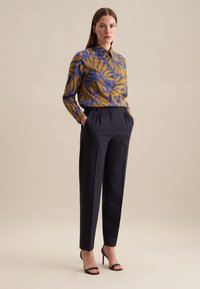 Camicia a maniche lunghe blu e senape con un motivo a foglie, abbinata a pantaloni scuri su misura e sandali neri con tacco. Tessuto della camicia liscio.