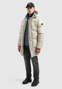 Gewatteerde lange jas in beige met een capuchon, ritssluiting en zijzakken. Draagt over een blauw shirt en donkere spijkerbroek, gecombineerd met zwarte schoenen.