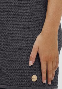 Dunkelgrauer gewebter Stoff mit strukturierter Oberfläche, versehen mit einem hölzernen Logo-Akzent am unteren Rand und einer Hand, die dagegen lehnt.