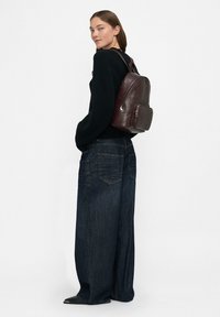 Donna che indossa un maglione nero, jeans a gamba larga scuri, scarpe nere e uno zaino in pelle marrone scuro, in piedi contro uno sfondo bianco semplice.