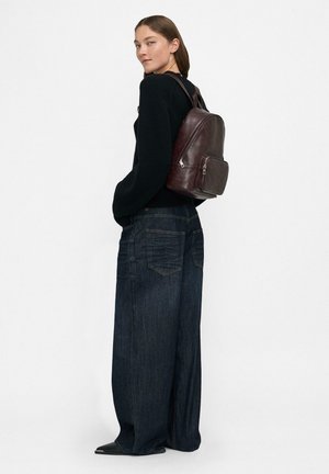 Donna che indossa un maglione nero, jeans a gamba larga scuri, scarpe nere e uno zaino in pelle marrone scuro, in piedi contro uno sfondo bianco semplice.