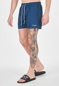 Bañador de color navy con cinturilla elástica y cordón blanco. Presenta un logotipo y una vista lateral de una pierna tatuada que lleva chanclas negras.