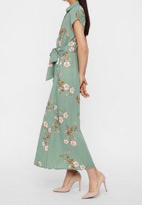 Robe florale vert clair avec des manches courtes, taille cintrée, et une silhouette fluide. Présente des motifs de fleurs blanches et roses sur l'ensemble.