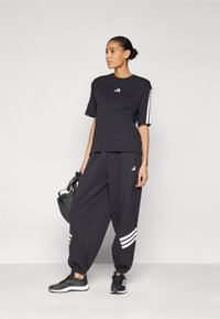 Schwarzes Sportoutfit mit einem locker sitzenden T-Shirt und Jogginghosen, beide mit weißen Akzentstreifen und Logodetails. Schwarze Sneaker runden das Erscheinungsbild ab.