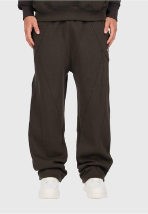 Homme portant un pantalon de survêtement ample marron foncé et des baskets blanches, se tenant debout avec les mains détendues sur les côtés sur un fond clair.