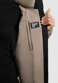 Chaqueta acolchada con un interior de color beige claro y un exterior negro, que presenta una etiqueta de parche y una textura suave. Mano visible sosteniendo el forro interior.