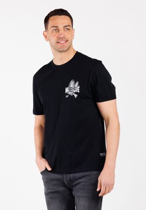 Hombre vestido con una camiseta negra que dice "Impresionante" y una gráfica de un águila, de pie con una mano en el bolsillo, mirando de lado, contra un fondo blanco.