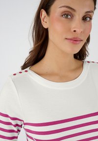 Witte top met roze horizontale strepen, ronde halslijn en drie decoratieve roze knopen op de schouder. Katoen materiaal, gladde textuur.
