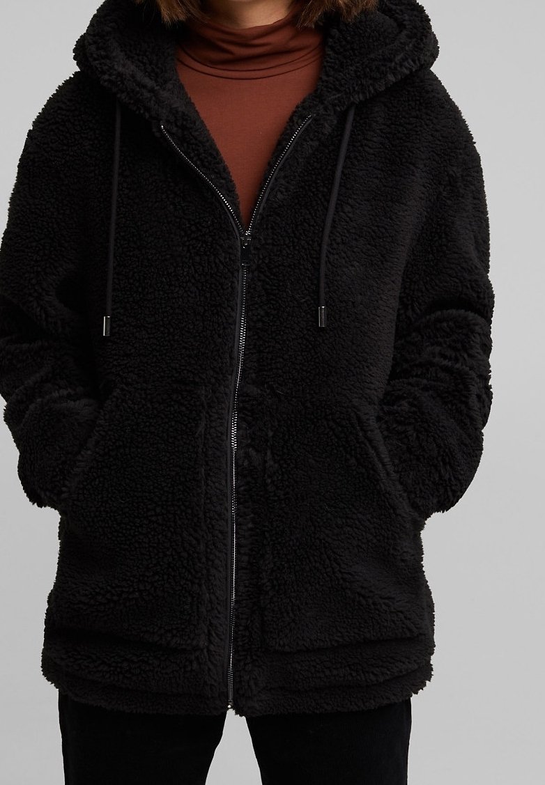 Personne portant une veste polaire sherpa noire zippée avec capuche et poches avant, par-dessus un col roulé marron.