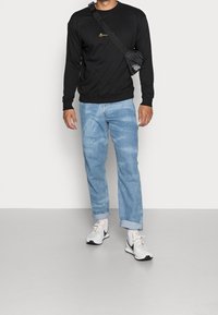 Zwarte trui met gouden borduursel, lichtblauwe spijkerbroek en witte sneakers. Inclusief een kleine zwarte crossbodytas over de schouder.