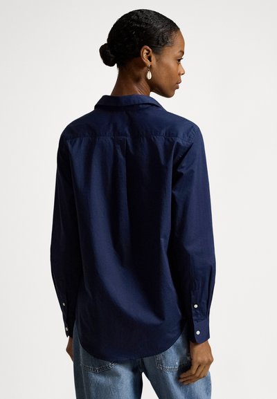 Ženska s temnimi lasmi v figi, oblečena v mornarsko modro majico z dolgimi rokavi in svetlo modre jeans hlače, obrnjena, da pokaže zadnji del majice.