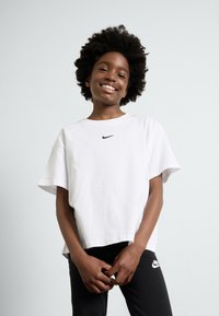 Biela bavlnená tričko s krátkymi rukávmi a čiernym logom Nike na hrudi, v kombinácii s čiernymi legínami s bielym logom na nohe.