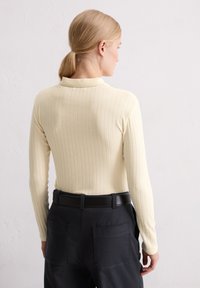 Frau mit blonden Haaren im Pferdeschwanz, die ein cremefarbenes, geripptes Langarmtop und schwarze Hosen mit einem schwarzen Gürtel trägt, steht vor einer weißen strukturierten Wand.