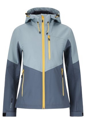 Persoon die een blauw en marineblauw jack met gele ritssluitingen draagt, zwarte broek en zwarte wandelschoenen, staat met gespreide benen.