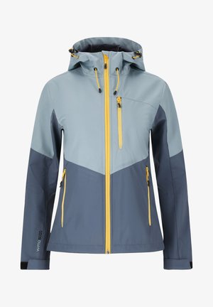 Personne portant une veste zippée bleue et marine avec des tirettes jaunes, un pantalon noir et des chaussures de randonnée noires, debout, jambes écartées.