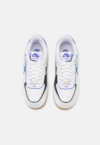 Bílé tenisky Nike Air Force 1 s černými akcenty, modrými detaily a perforovanou špičkou. Klasický design, gumová podrážka pro dobrou přilnavost.