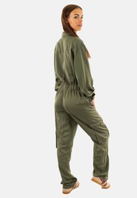 Jumpsuit verde oliva con vita regolabile, maniche lunghe, tasche frontali e cerniere sulle gambe. Tessuto liscio e strutturato.