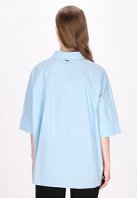 Hellblaues, übergroßes Hemd mit kurzen Ärmeln, Knopfleiste und naht in der Mitte des Rückens. Glattes, leichtes Material, minimalistisches Design.
