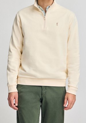 Ljusbeige pullover-tröja med halvdragkedja vid kragen, mjuk textur och en liten broderad logotyp på bröstet. Bärs med gröna byxor.