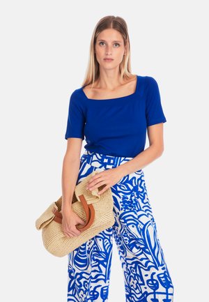 Blue short-sleeve top with a square neckline, paired with patterned white and blue wide-leg pants. Holding a woven straw bag with leather accents.