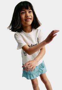 Kortärmad krämfärgad t-shirt med svart brodyr, kombinerad med en ljusblå volangkjol. Barnet ler och poserar med utsträckta armar.