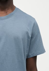 Άνδρας που φοράει απλό, ανοιχτό μπλε κοντομάνικο T-shirt με λευκό φόντο, κομμένο στους ώμους και στο πάνω μέρος του στήθους.