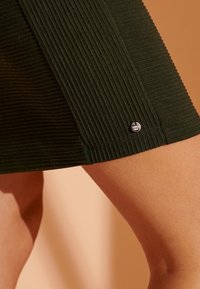 Grön ribbad kjol i ett texturerat tyg, med en liten silverlogotyp vid fållen. Slät yta med en strukturerad silhuett.