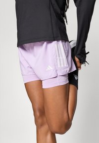 Shorts de sport lilas clair avec doublure intérieure, ornés de trois rayures blanches et d'un logo Adidas. Le tissu a une texture lisse et légèrement brillante.