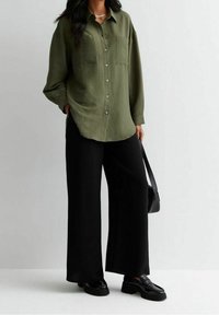 Kvinna som bär en olivgrön knäppt skjorta, svarta vida byxor, svarta loafers och håller en svart handväska framför en enkel bakgrund.