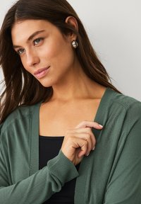 Cardigan verde realizzato in tessuto morbido con finiture a costine, indossato sopra una maglietta nera. Il modello tocca il cardigan, evidenziando la sua texture e vestibilità.