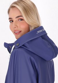 Fialová nepremokavá bunda s vysokým golierom, bielym šnúrkou a kontrastným kovovým príslušenstvom; hladká textúra a jemný loga detail na kapucni.