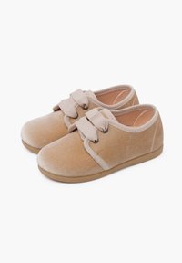 Scarpe in velluto beige con punta arrotondata, fasce elastiche e due fiocchi decorativi sulla parte anteriore. Materiale morbido e suola piatta in gomma.