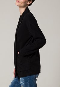 Giacca leggera nera con chiusura a bottoni, tasche laterali e vestibilità rilassata, abbinata a jeans blu strappati. Tessuto morbido.