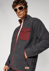 Sötét szürke fleece dzseki bordó cipzáras zsebbel, bordás gallérral és hímzett logóval. Az anyag textúrájának látszódnia kell, és puha.