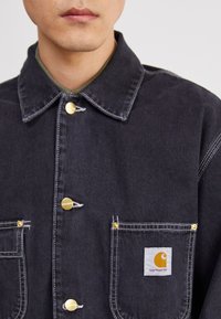 Dark denim jacket featuring a collar, gold buttons, chest pockets, and a Carhartt logo patch. Contrast stitching adds detail.