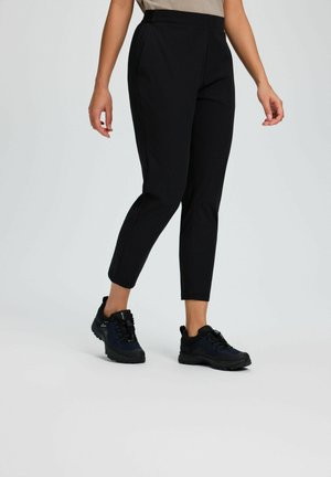 Person trägt schwarze, kurz geschnittene Hosen und schwarze Wanderschuhe, steht vor einem einfarbigen hellgrauen Hintergrund.