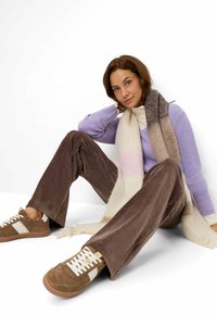 Paarse gebreide trui, bruine corduroy broek en bruine sneakers met witte accenten. Een zachte, gestreepte sjaal hangt om de nek.