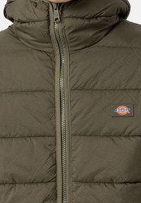Kurtka puchowa w oliwkowej zieleni z wysokim kołnierzem, pełnym suwakiem, pikowanym wzorem i okrągłą naszywką z logo na piersi.