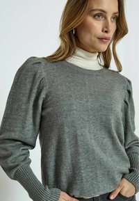 Pilkos megztos palaidinės su pūstomis rankovėmis ir raukšlėtais manžetais. Lygus paviršius ir apvalus kaklas. Dėvima ant kreminės turtlenecko.