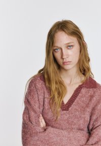 Mehak pleten pulover v svetlo rdeči barvi z globokim V-izrezom in kontrastnim ovratnikom. Ima široke rokave in teksturirano površino.