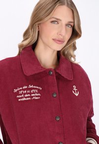 Giacca in velluto a coste bordeaux con un ampio colletto, bottoni neri e testo ricamato. Presenta un motivo ad ancora sul lato.