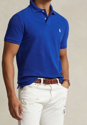 Polo à manches courtes bleu roi avec une texture tricotée et une patte de boutonnage à trois boutons. Porté avec un jean blanc déchiré et une ceinture tressée marron.